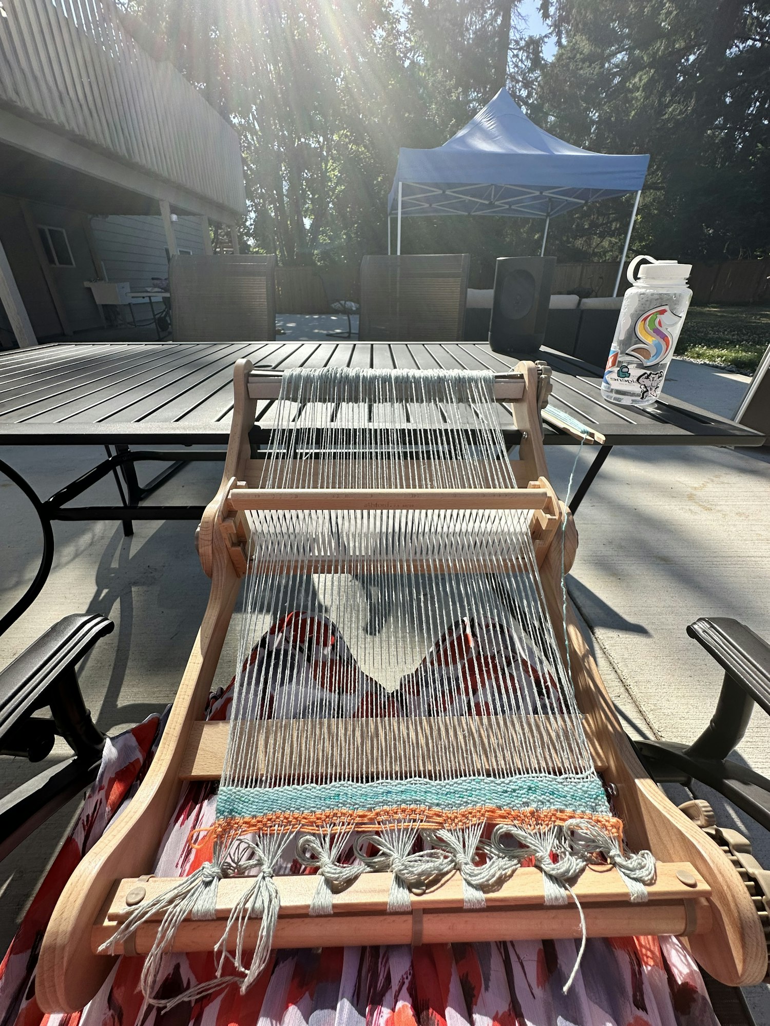 Arisa outside on a sunny day with her Ashford Knitter's Loom on her lap. There's green-blue weft yarn, gray-blue warp yarn, and orange scrap yarn.