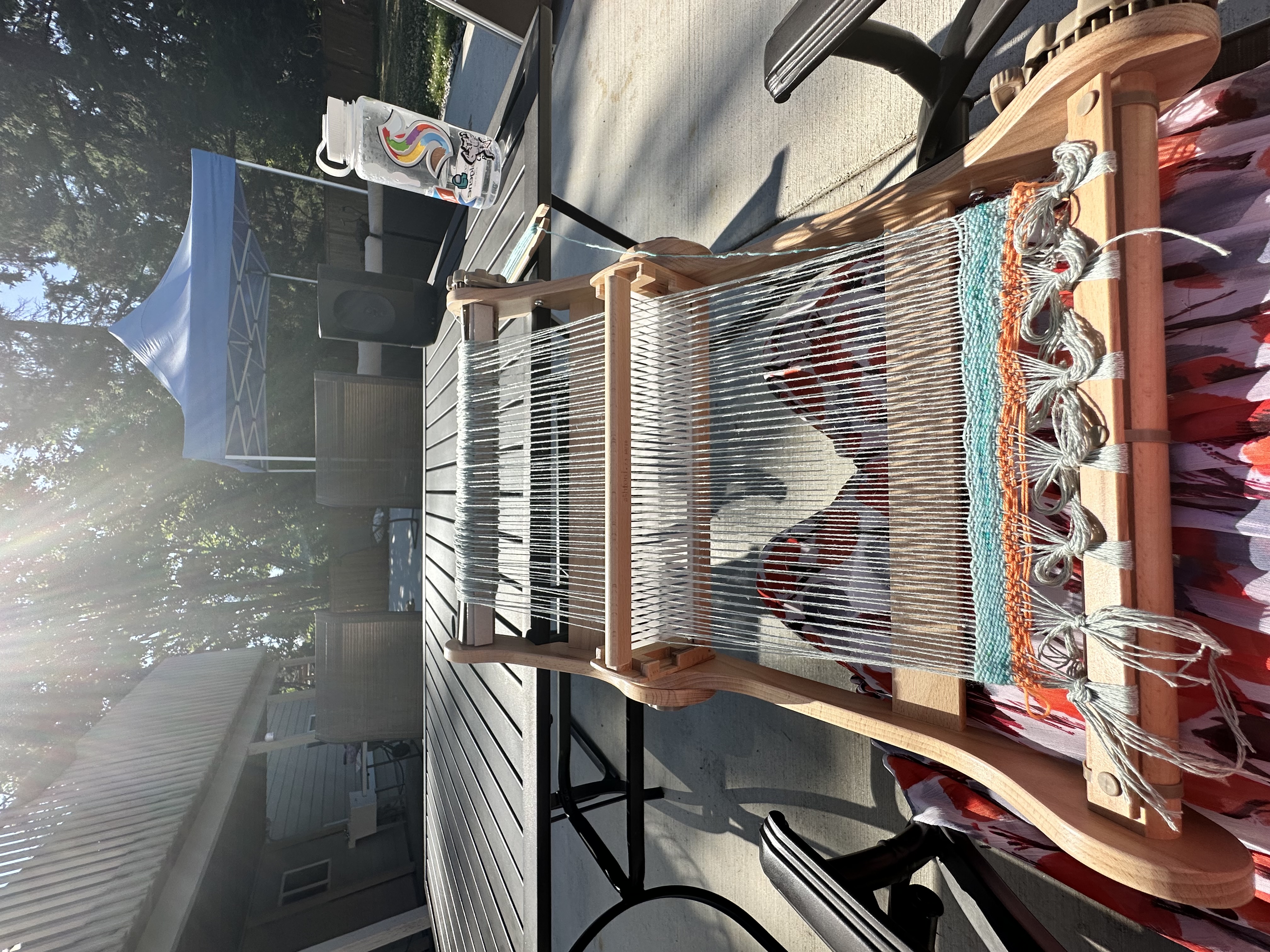 Arisa outside on a sunny day with her Ashford Knitter's Loom on her lap. There's green-blue weft yarn, gray-blue warp yarn, and orange scrap yarn.