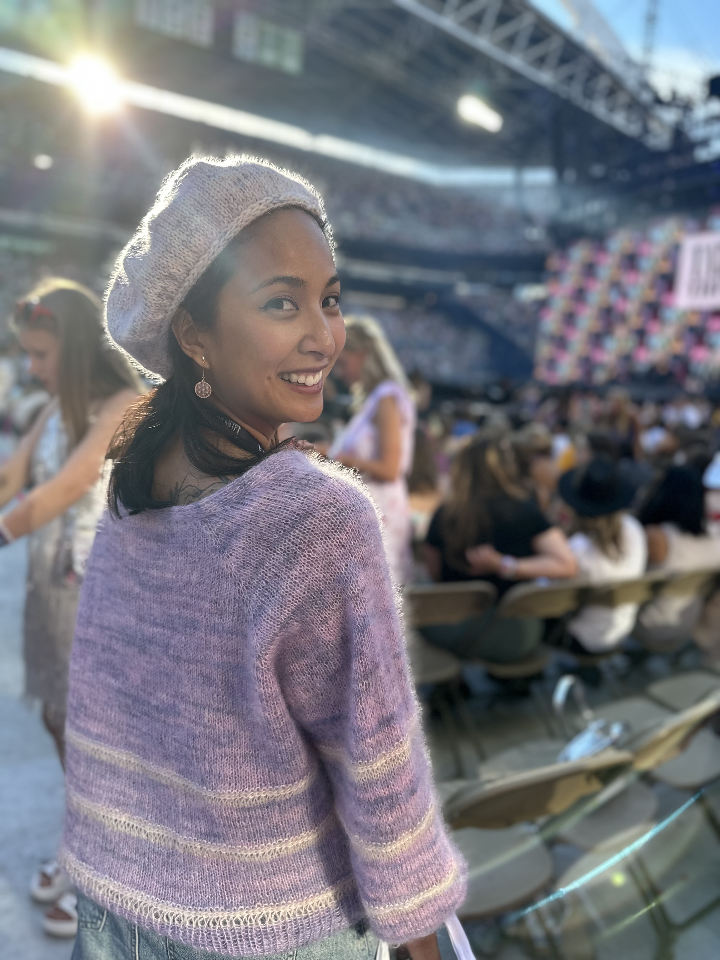 Arisa from behind with her face smiling at the camera. She's where a beret and cardigan in pink, purple, and white at the Taylor Swift concert